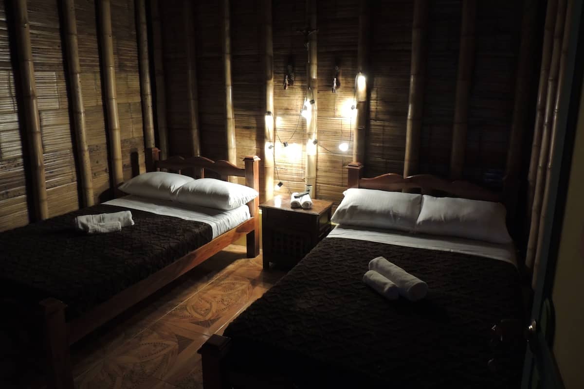 Two single beds with wooden frames and headboards, a nightstand with a lamp, and a wall with vertical bamboo slats.