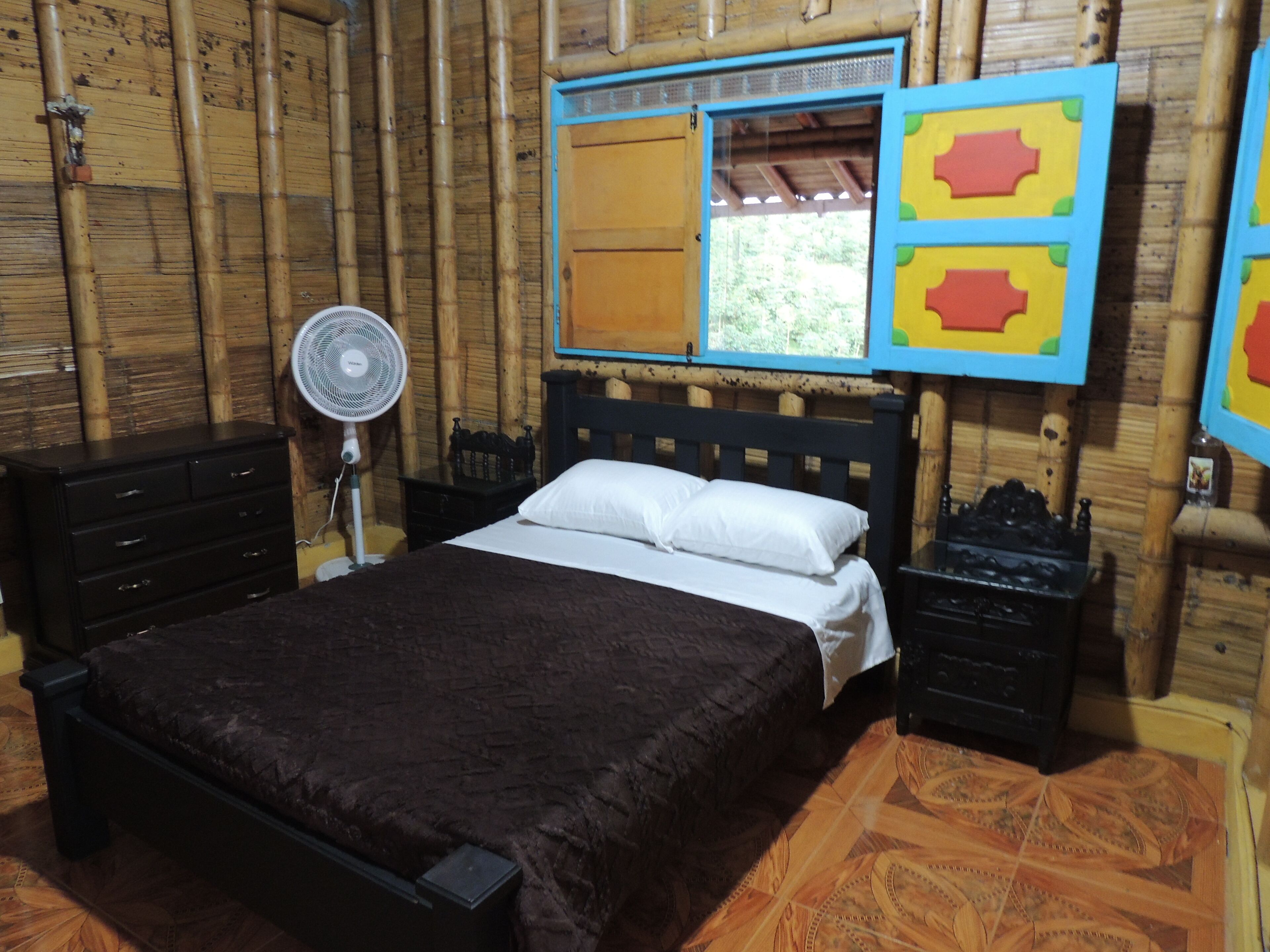 A bedroom with a bed, a nightstand, a dresser, a fan, and a window with a view of greenery.