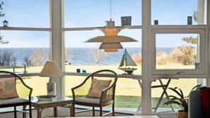 A room with large windows overlooking a beach, featuring two wicker chairs, a table, and a lamp. - Awesome home in Sjællands Odde (Sjællands Odde)