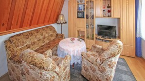 Uma sala de estar aconchegante com um sofá estampado, duas poltronas, uma mesa redonda pequena, uma televisão e armários de madeira com portas de vidro.