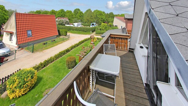 Varanda com vista para um jardim, um deck de madeira e um edifício com telhado vermelho ao fundo.