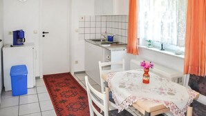 Cocina compacta con refrigerador, cafetera y lavamanos. Hay una mesa de comedor con mantel floreado y un jarrón de flores. Se ven cortinas naranjas y una ventana con vistas.