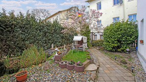 Ein Garten mit gepflastertem Weg, einer kleinen Sitzecke und verschiedenen Pflanzen und Bäumen.