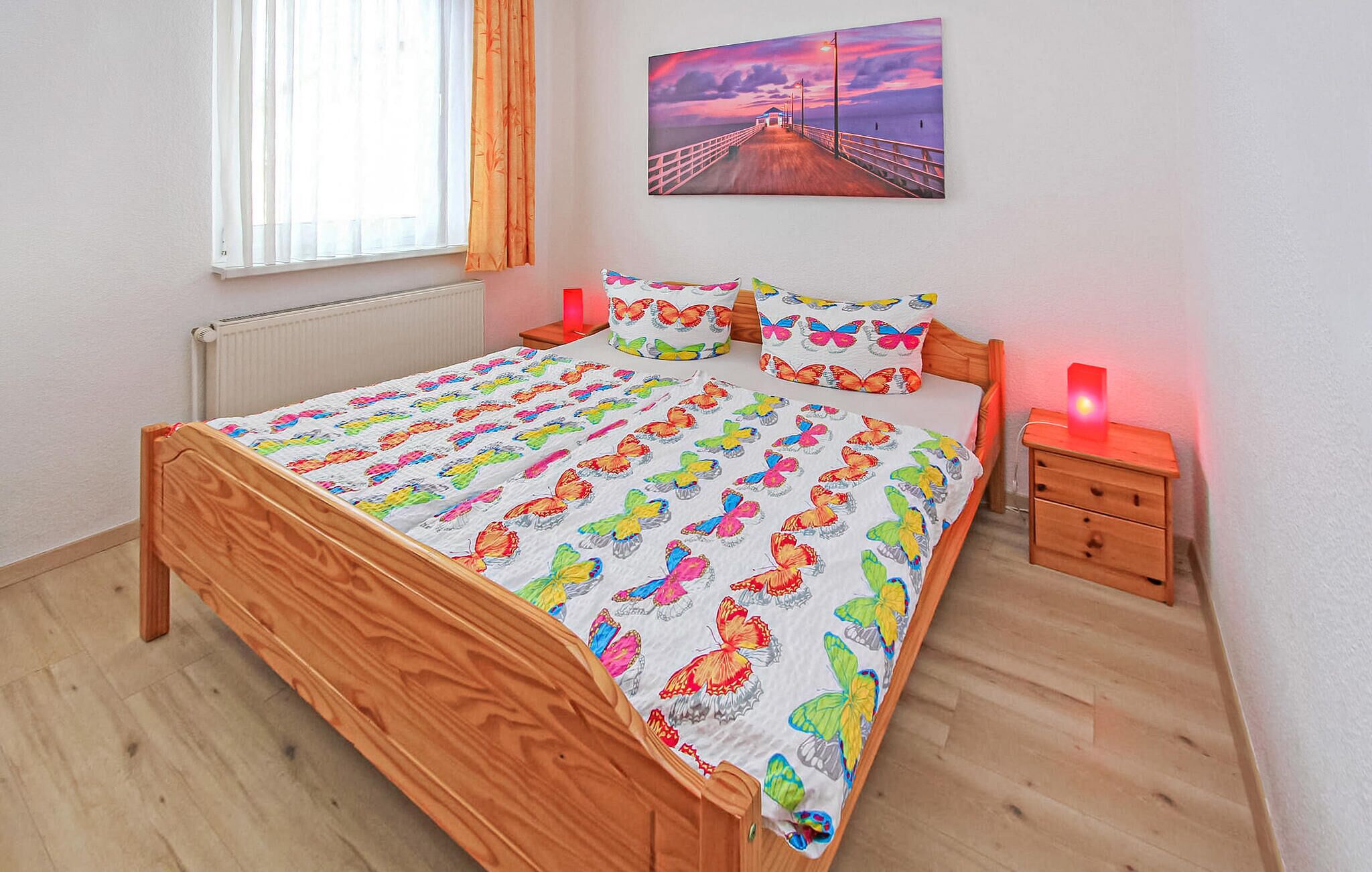 A bedroom with a wooden bed, colorful butterfly bedding, a nightstand with a lamp, and a painting of a pier on the wall.