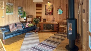 Una habitación acogedora con un sofá de cuero negro, suelo de madera, un tapete grande, una pared de madera adornada con cuadros y una lámpara colgante, además de un estufa de leña negra.