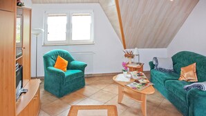 Un salon chaleureux avec un plafond haut, un meuble en bois, un fauteuil turquoise agrémenté d’un coussin orange, une table basse en bois sur laquelle est posée une courtepointe, et un canapé vert avec un coussin orange.