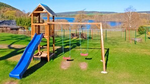 A playground with a slide and swings, set against a backdrop of a lake and hills.