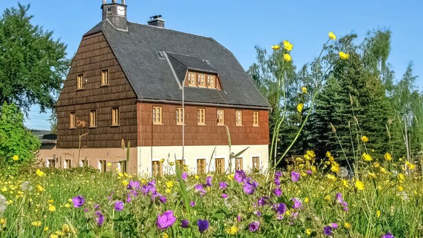 A traditional wooden building with a steep gabled roof and a bell tower, surrounded by a field of wildflowers.