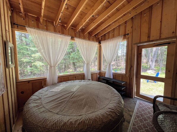 A wooden cabin bedroom with a round bed, two windows with sheer curtains, a small table, and a chair. - Relaxing tranquil setting, hot tub, fire pit; Pet friendly. (Middle Grove)