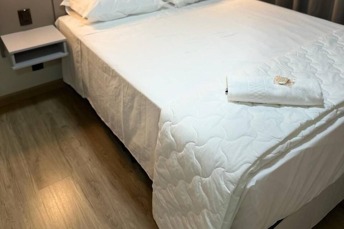 Una cama bien hecha con sábanas blancas, una mesita de noche con una lámpara y un suelo de madera.