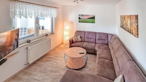 Un salon moderne avec un canapé d’angle, une table basse ronde, une grande fenêtre offrant une belle vue et un tableau accroché au mur.