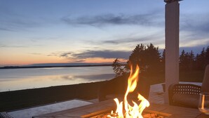 Feuerstelle mit Flammen, umgeben von Gartenmöbeln, mit Blick auf ruhiges Gewässer und einen bewaldeten Bereich bei Sonnenuntergang.