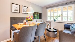 A modern dining area with a wooden table set for four, grey upholstered chairs, a grey sofa, and a large window with blinds. - Watt. Familie. Reet. - Erich Johannsen Wai (Keitum)