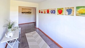 A hallway with a tiled floor, a white wall with fruit paintings, a small table with a teapot, and a potted plant. - Ehemaliges Farmhaus Lehsten (Lehsten)