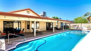A swimming pool with a patio area, outdoor seating, and a building with a red roof. - Sunny Oasis 4BR Pool Home with Game Room (Mesa)
