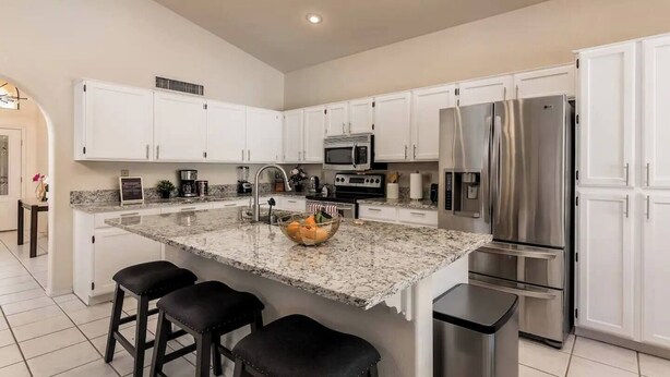 A modern kitchen with white cabinets, stainless steel appliances, a granite countertop, and a central island with stools. - Sunny Oasis 4BR Pool Home with Game Room (Mesa)