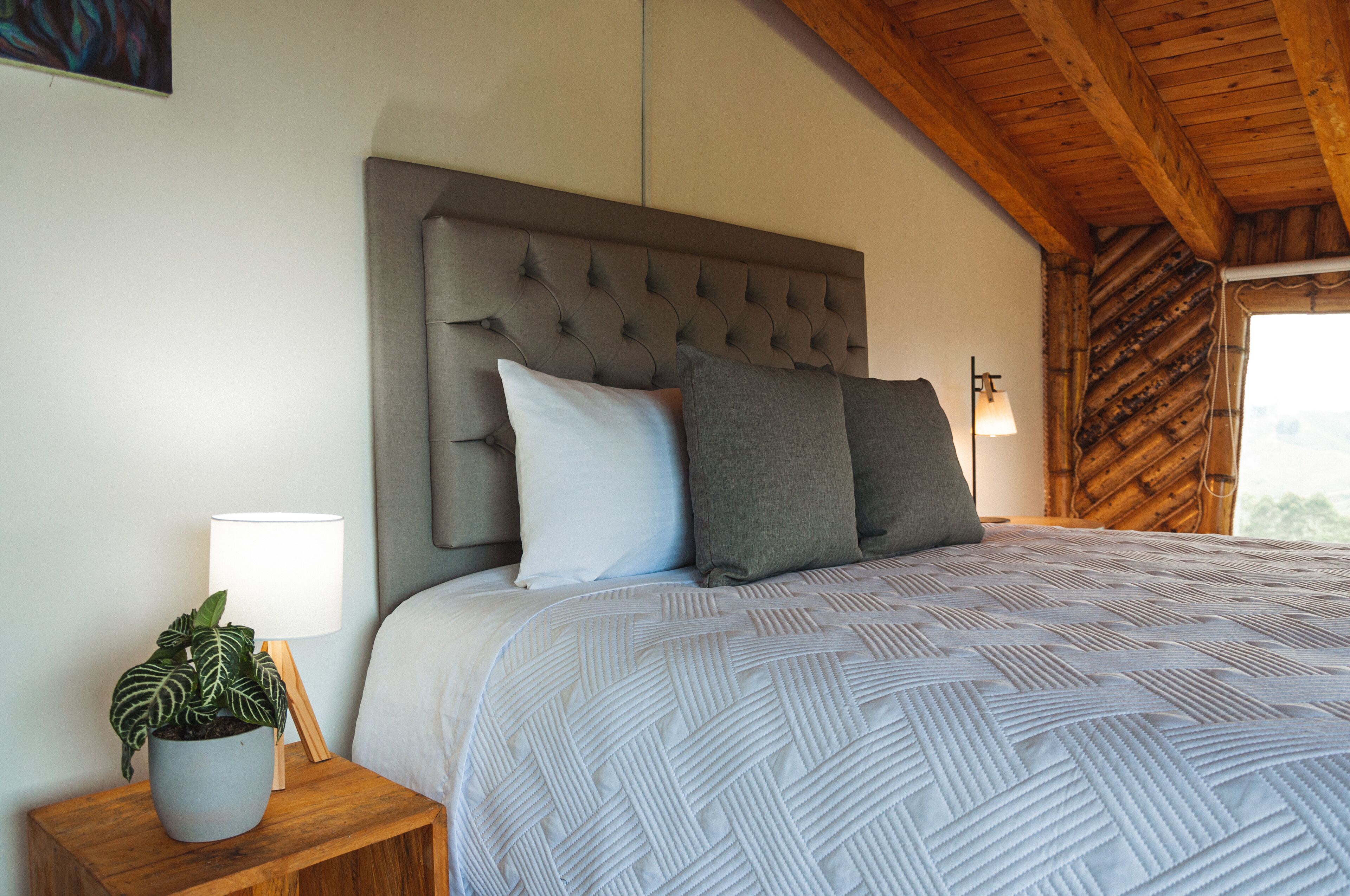 Une chambre à coucher avec une tête de lit capitonnée, un lit recouvert d’une couvre-lit à motifs, une table de chevet avec une lampe et une plante en pot, et un plafond en bois.