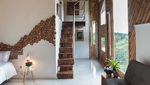 A staircase made of wooden logs, a bed, a sofa, a small table with a lamp, and a view of greenery through large windows. - Hotel PIA Colombia (Marinilla)