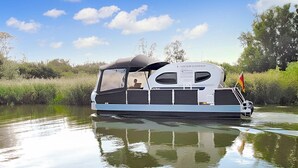 A riverboat with a covered deck and a flag, moving through a calm river with greenery on the banks. - Nice ship in Verchen with kitchen (Verchen)