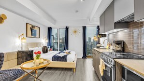 A modern hotel room with a bed, a sofa, a small dining table, a kitchen area with a stove, and a view of the city through large windows. - Panorama Suites (Toronto)