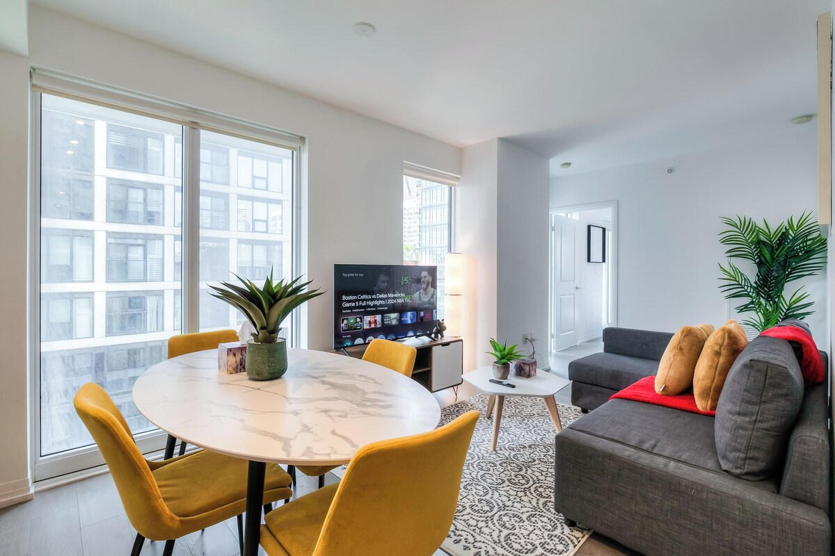 Una sala de estar moderna con una mesa de comedor, sillas amarillas, un sofá gris y un televisor.