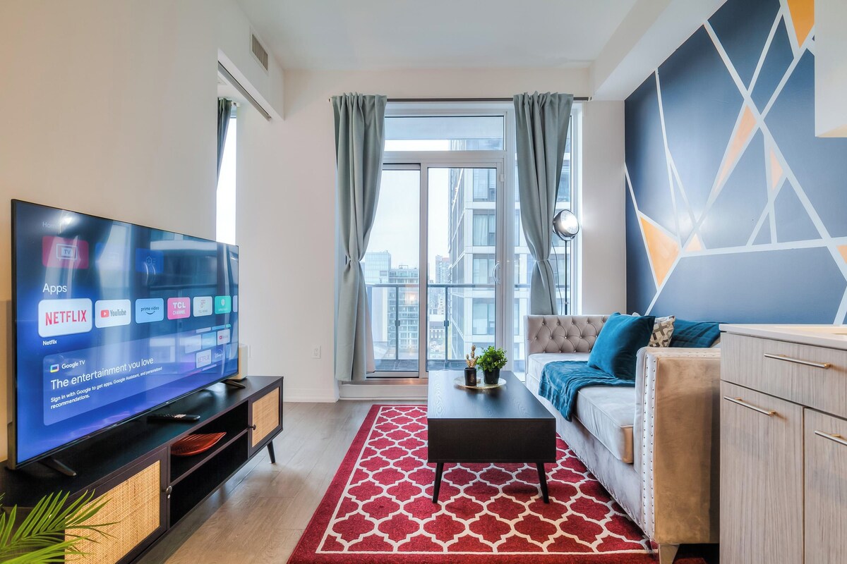 Una sala de estar moderna con televisor de pantalla plana, sofá, una mesita de centro y vistas de la ciudad a través de amplios ventanales.