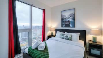 A hotel room with a large bed, two bedside lamps, a nightstand, a green throw blanket, and a window with red curtains.