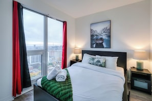 A hotel room with a large bed, two bedside lamps, a nightstand, a green throw blanket, and a window with red curtains. - Polaris Suites (Toronto)