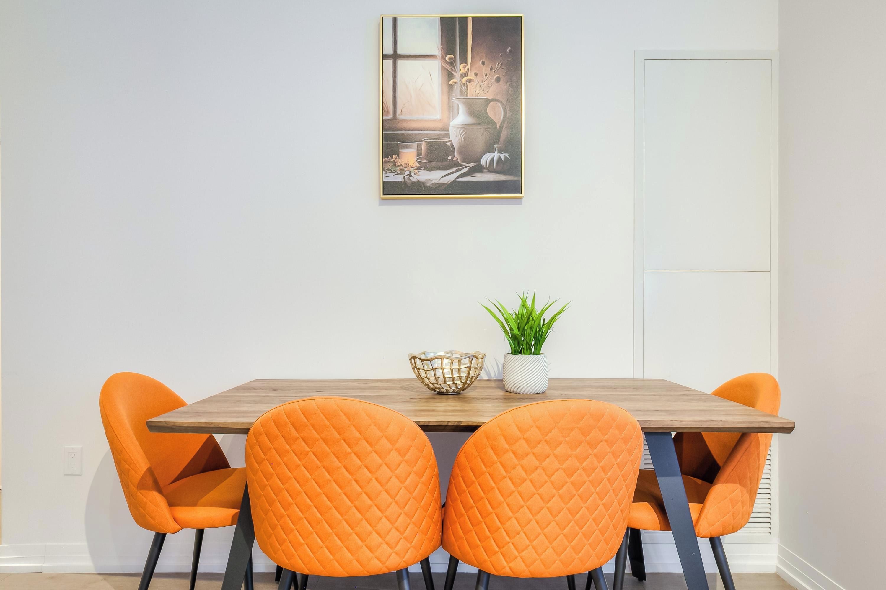 Ruokailutilassa on puinen pöytä, neljä oranssia tuolia, maljakko kukkineen ja seinällä oleva kehystetty kuva.