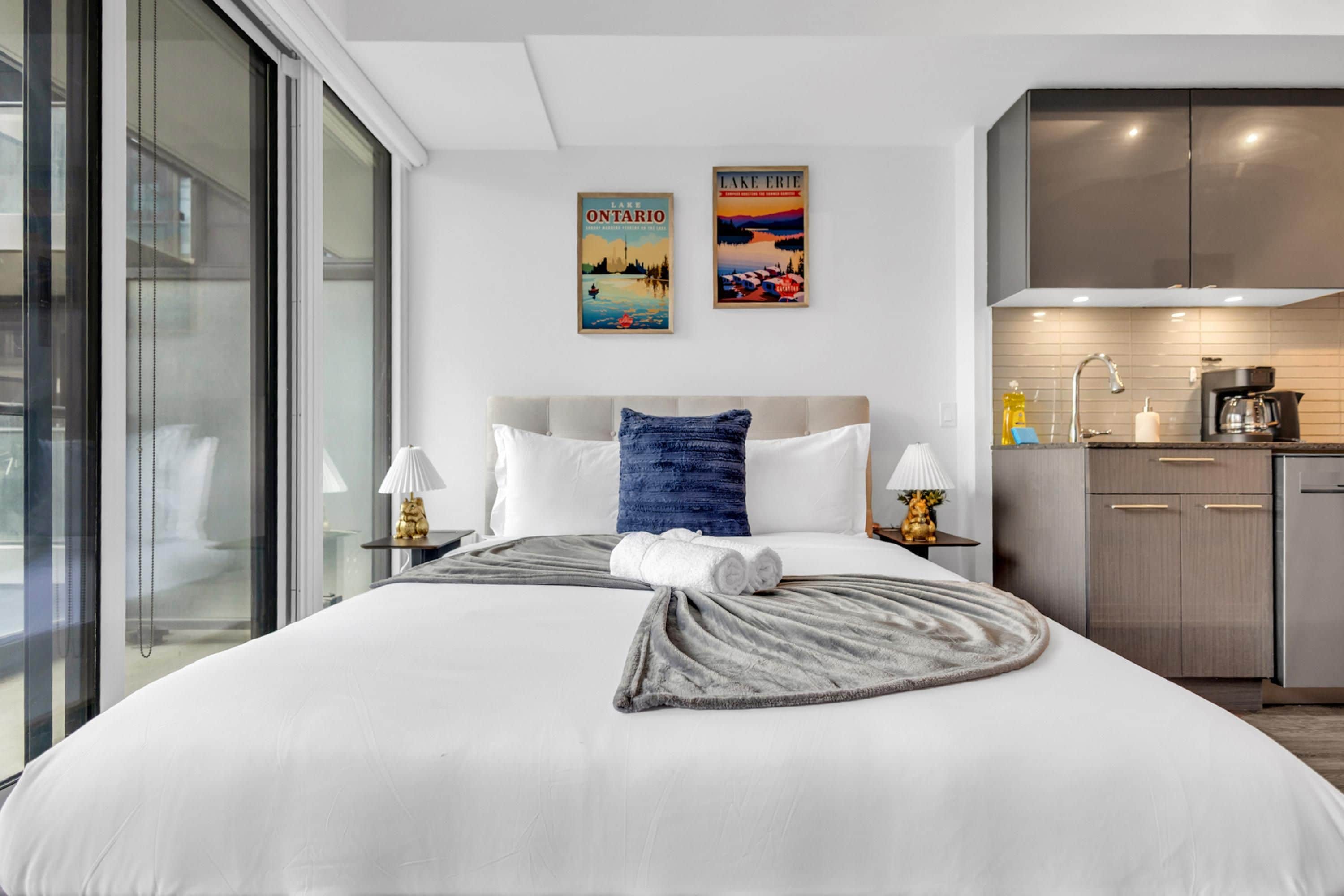 Une chambre d’hôtel moderne dotée d’un grand lit, offrant une vue sur la ville, d’une kitchenette et de deux œuvres d’art murales.