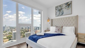 A bedroom with a large window, a bed with white linens, a blue throw blanket, and a city view. - Panorama Suites (Toronto)