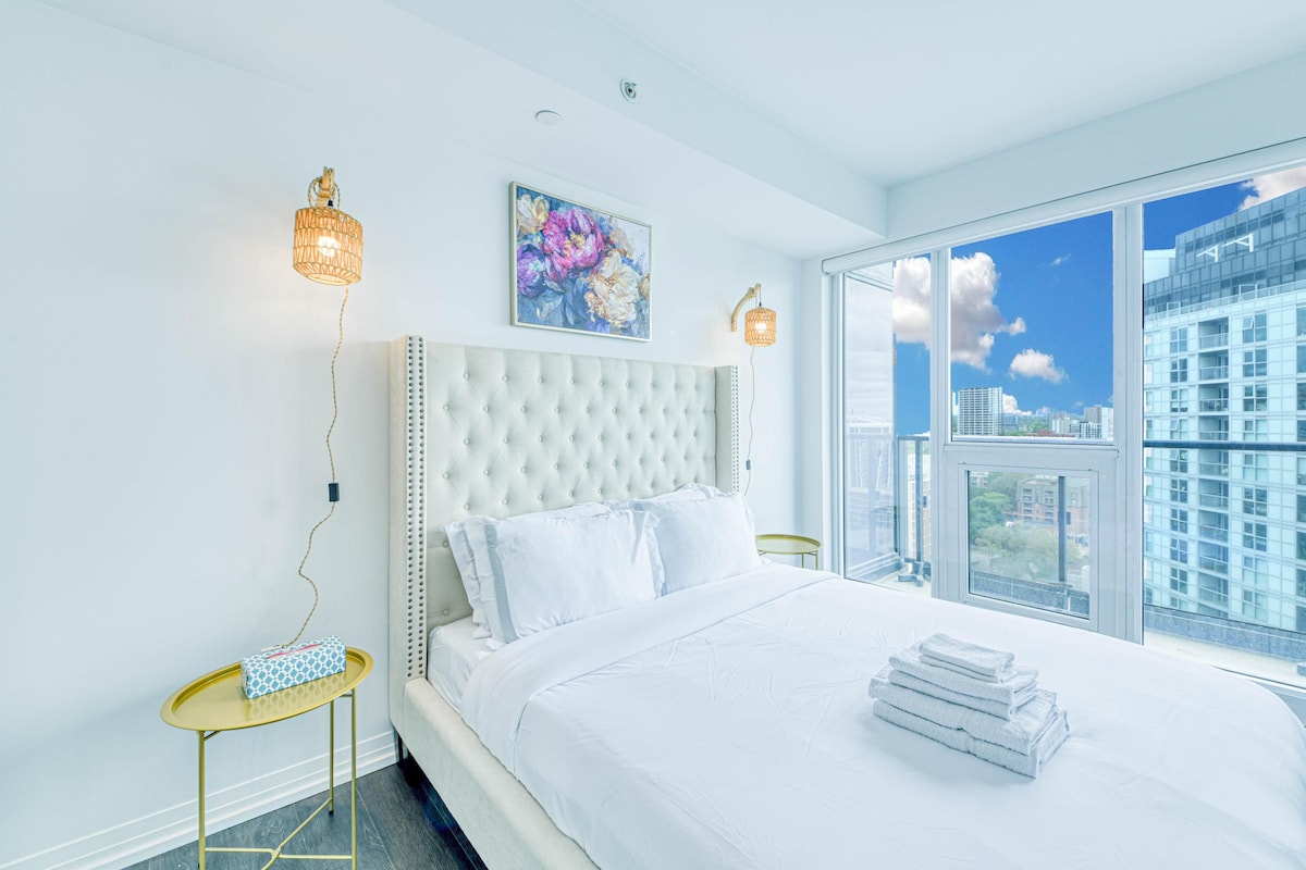 Un dormitorio con cabecera capitonada, un ventanal con vistas a la ciudad, una mesita de noche con lámpara y una cama bien hecha.