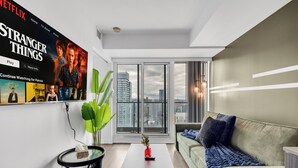 A modern living room with a large window, a green sofa, a coffee table, and a wall-mounted TV displaying 'Stranger Things'. - Panorama Suites (Toronto)