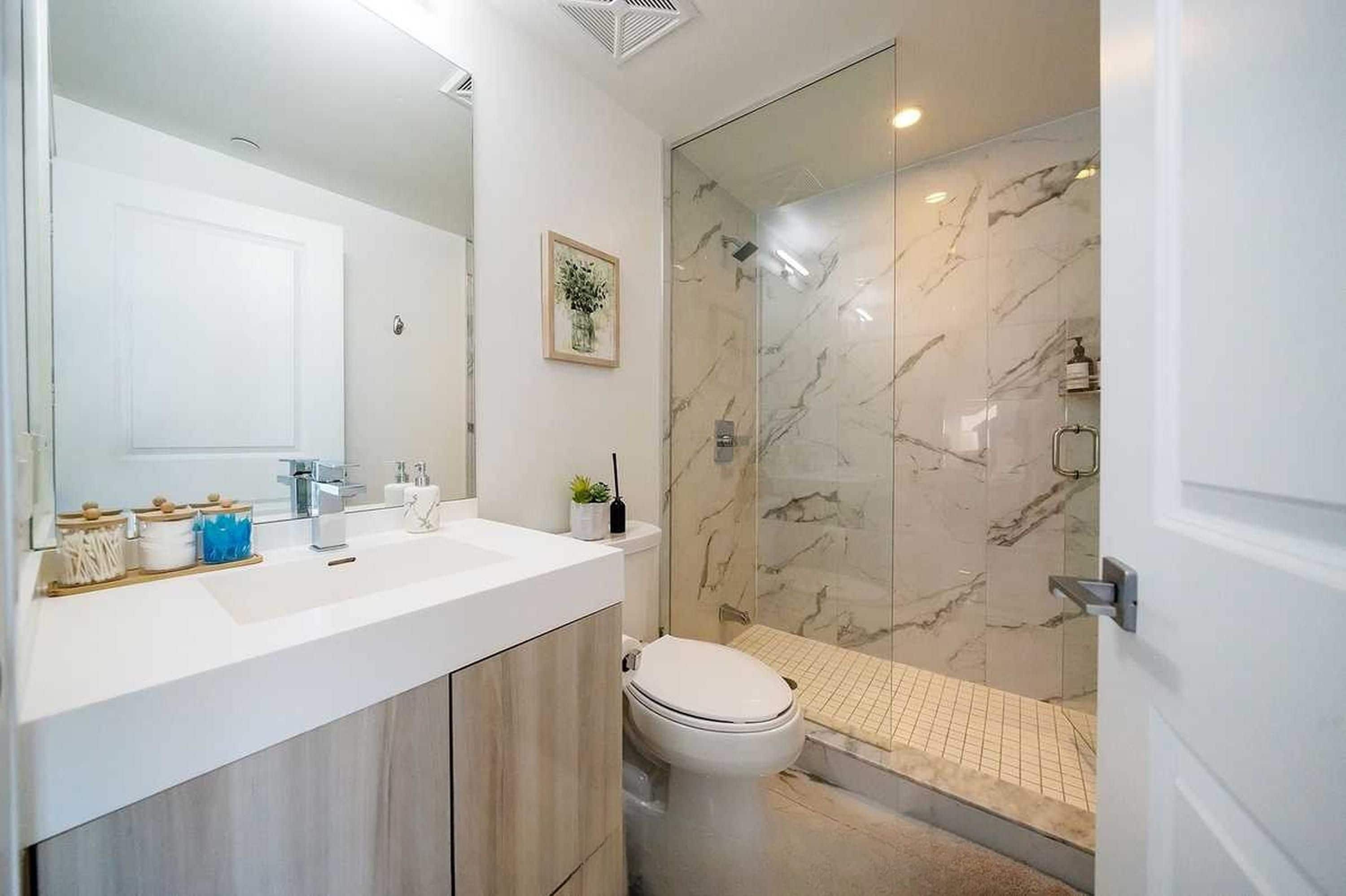 A modern bathroom with a white sink, mirror, and toilet. It features a glass shower enclosure with marble walls and a tiled shower floor.