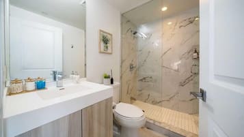 A modern bathroom with a white sink, mirror, and toilet. It features a glass shower enclosure with marble walls and a tiled shower floor.