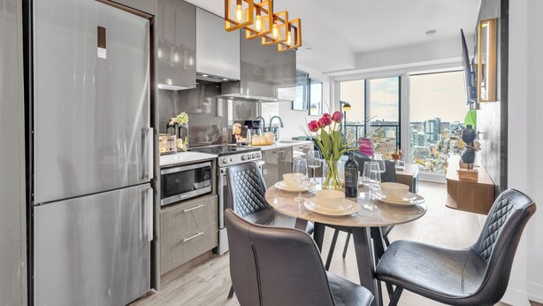 A modern kitchen with stainless steel appliances, a dining area with a round table, and a city view through large windows. - Panorama Suites (Toronto)
