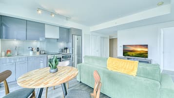 A modern kitchen with grey cabinets, a dining table with chairs, a green sofa with a yellow throw, and a TV displaying a sunset.