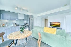 A modern kitchen with grey cabinets, a dining table with chairs, a green sofa with a yellow throw, and a TV displaying a sunset. - Polaris Suites (Toronto)