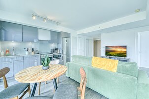 Une cuisine moderne avec des meubles gris, une table à manger avec des chaises, un canapé vert agrémenté d’un plaid jaune et un téléviseur diffusant une image de coucher de soleil.