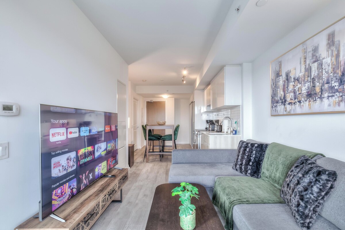 Una sala de estar moderna con un televisor de pantalla plana mostrando la interfaz de un servicio de streaming, un sofá gris con almohadones decorativos, una mesa de centro de madera con una planta y un cuadro de un paisaje urbano en la pared.