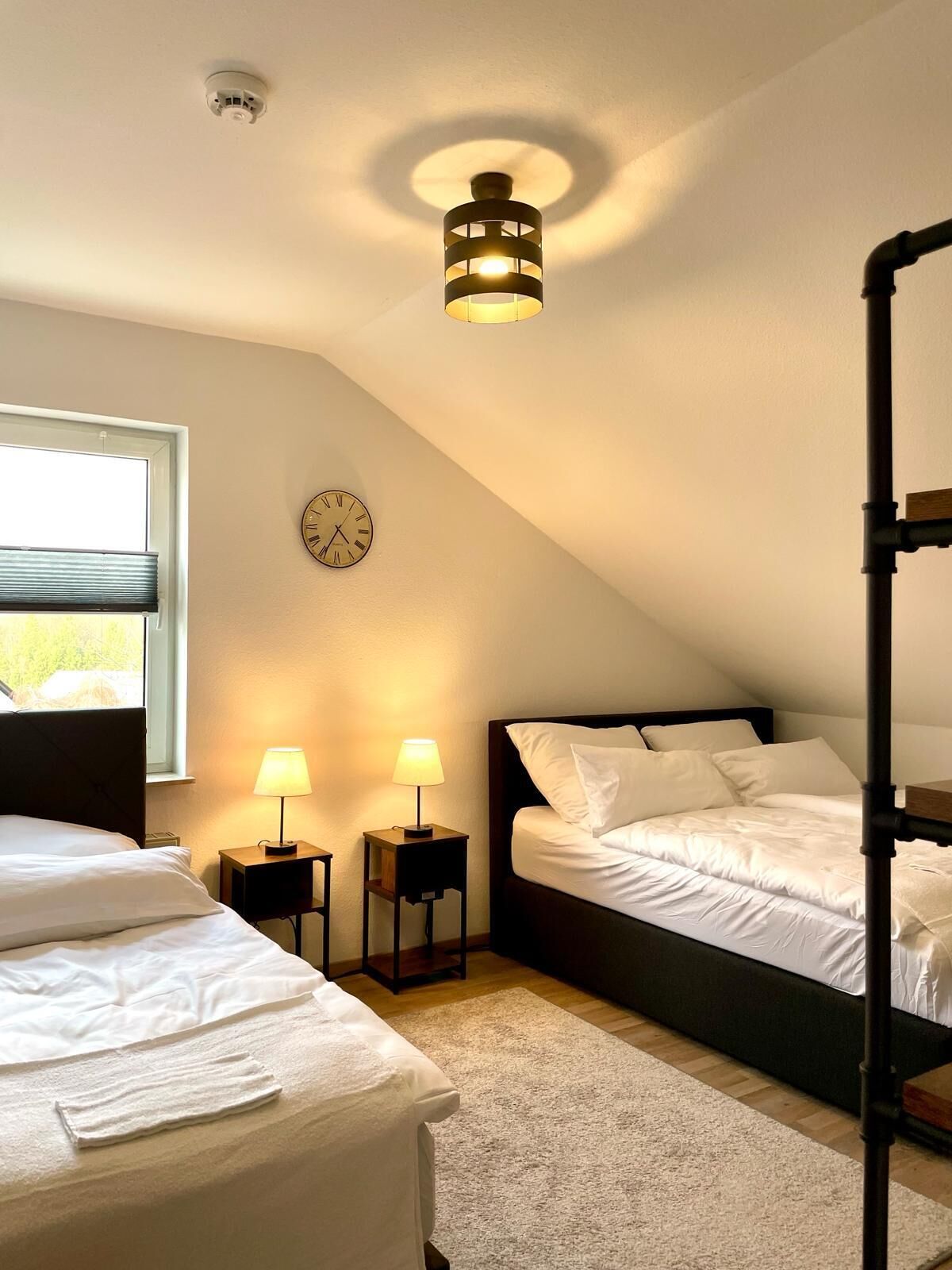 Quarto com duas camas, uma luminária no teto, um relógio e uma janela com persianas.