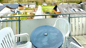 A balcony with a table and chairs, overlooking a residential area with houses and a forested hill.