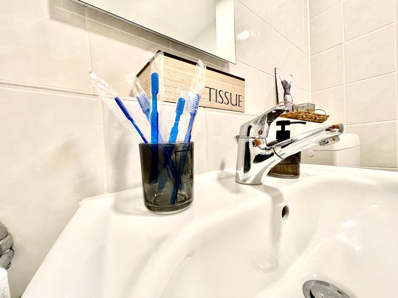 Un lavamanos con un porta cepillos de dientes que contiene cepillos azules, un dispensador de jabón y una caja de pañuelos en la pared con la etiqueta 'TISSUE'.