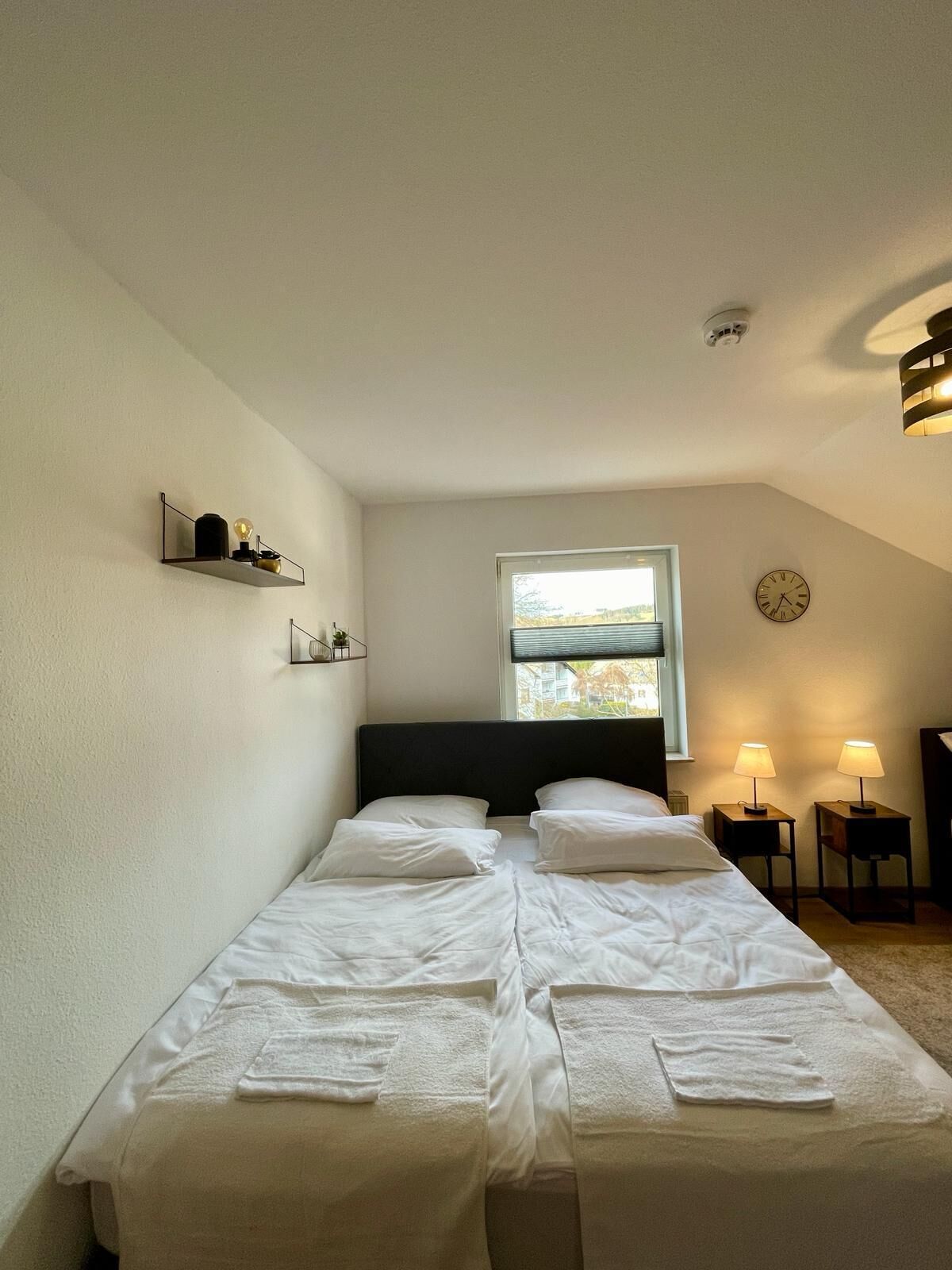 Une chambre à coucher comprenant un lit, deux tables de chevet, une fenêtre, une horloge murale et une étagère avec des objets décoratifs.