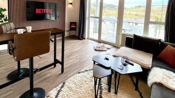 A modern living room with a wooden ceiling, a large TV screen displaying 'NETFLIX', a cozy seating area with a sofa and armchairs, a coffee table, and a fireplace.