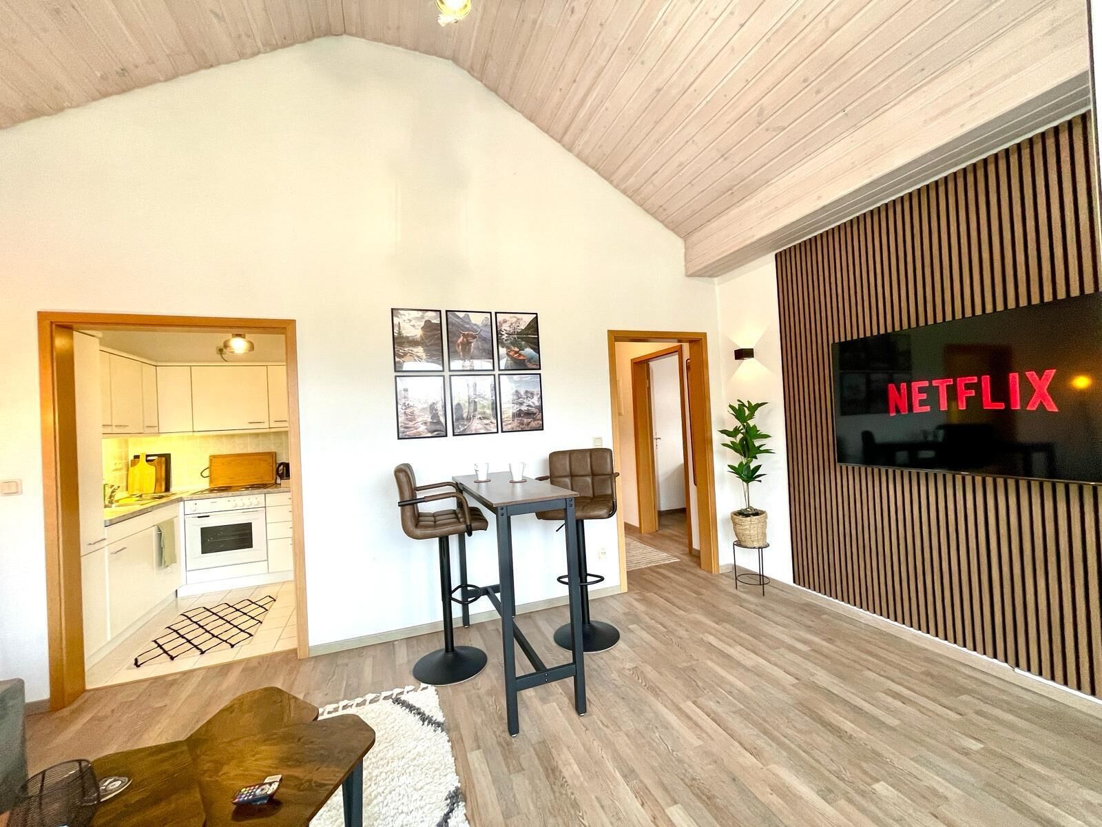 Ein modernes Wohnzimmer mit Küche im Hintergrund, einer Essecke mit Hockern und einem großen Fernsehbildschirm, der "NETFLIX" anzeigt.