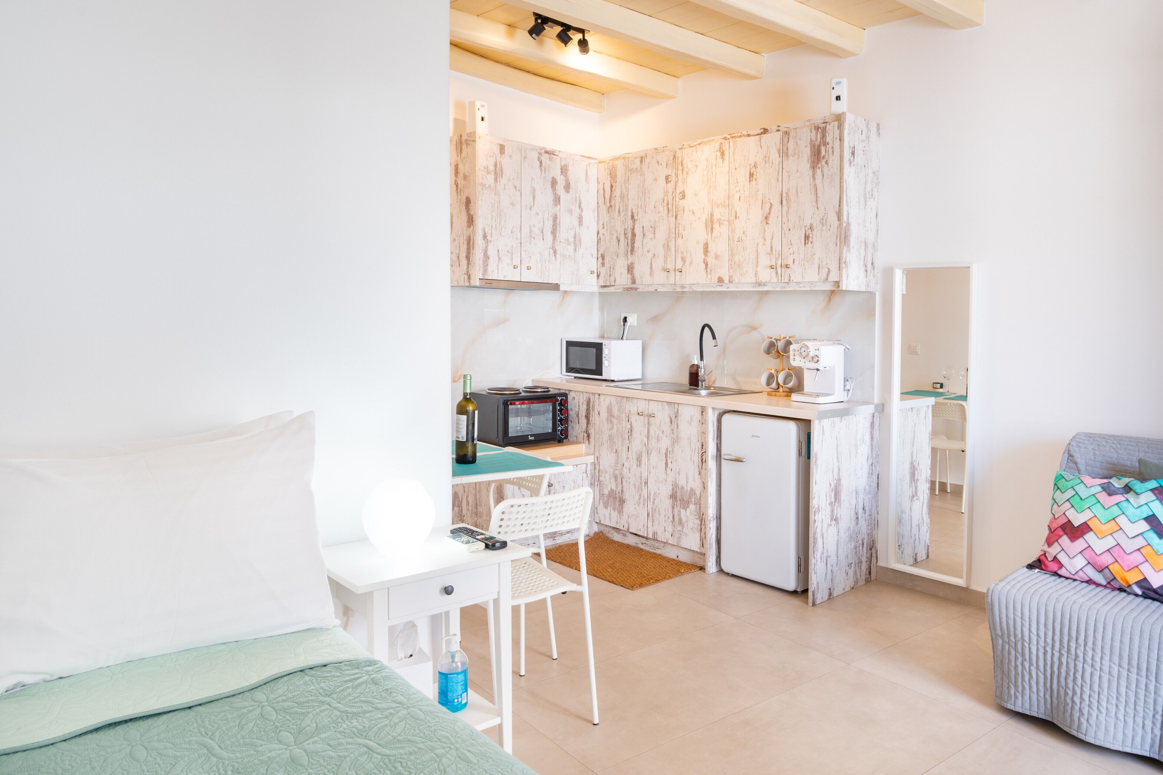 Een kleine keuken met een magnetron, broodrooster en koelkast, een eethoekje met een stoel en een bed met een groene deken.