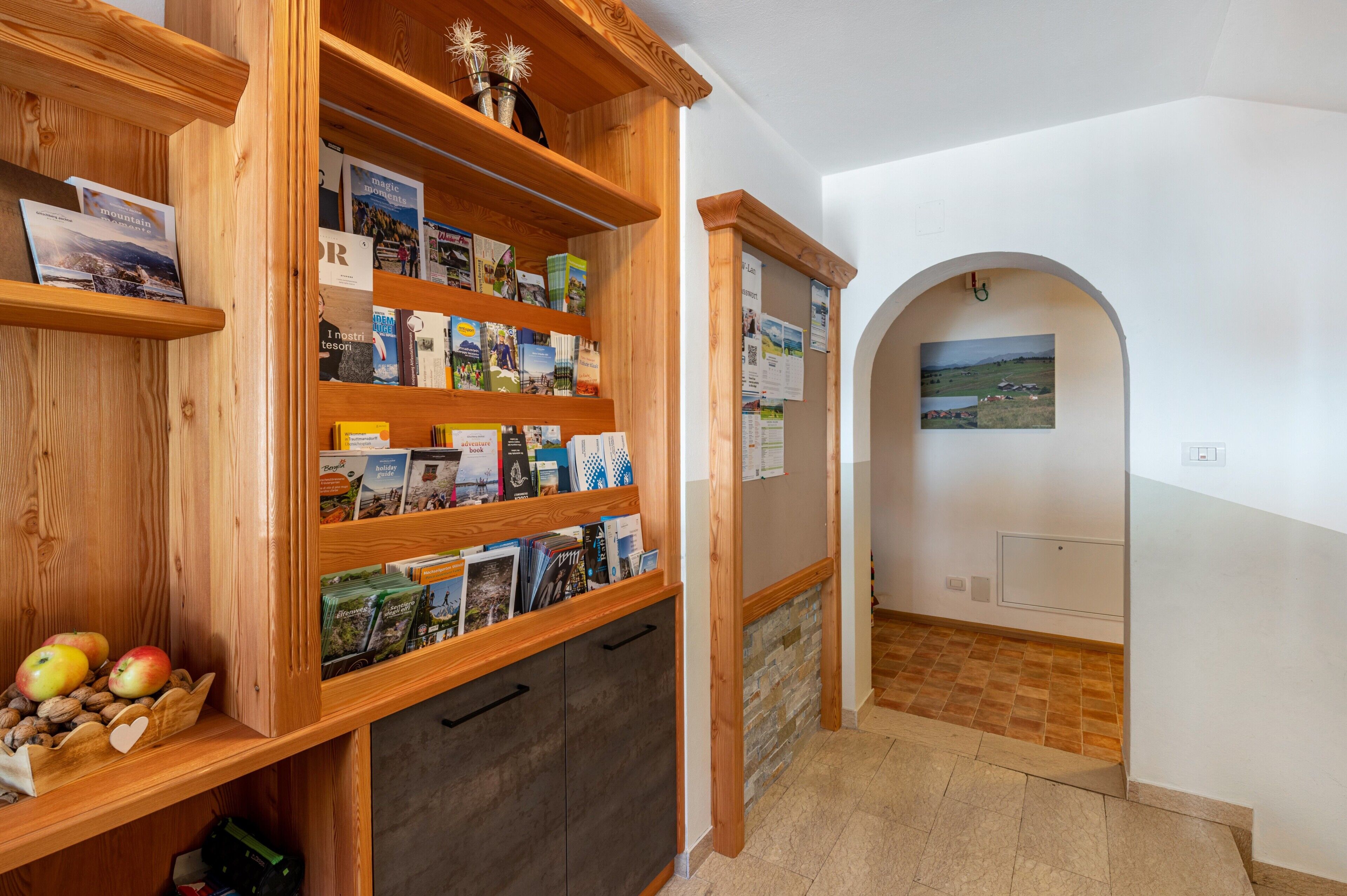 A wooden bookshelf with travel brochures, a small display of apples and nuts, and a view through an archway to a landscape.