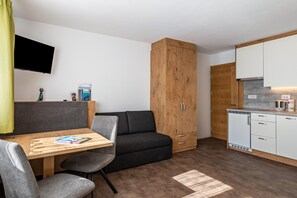 A compact room with a sofa, a small table with a magazine, a wooden cabinet, a TV mounted on the wall, and a kitchenette with a dishwasher.