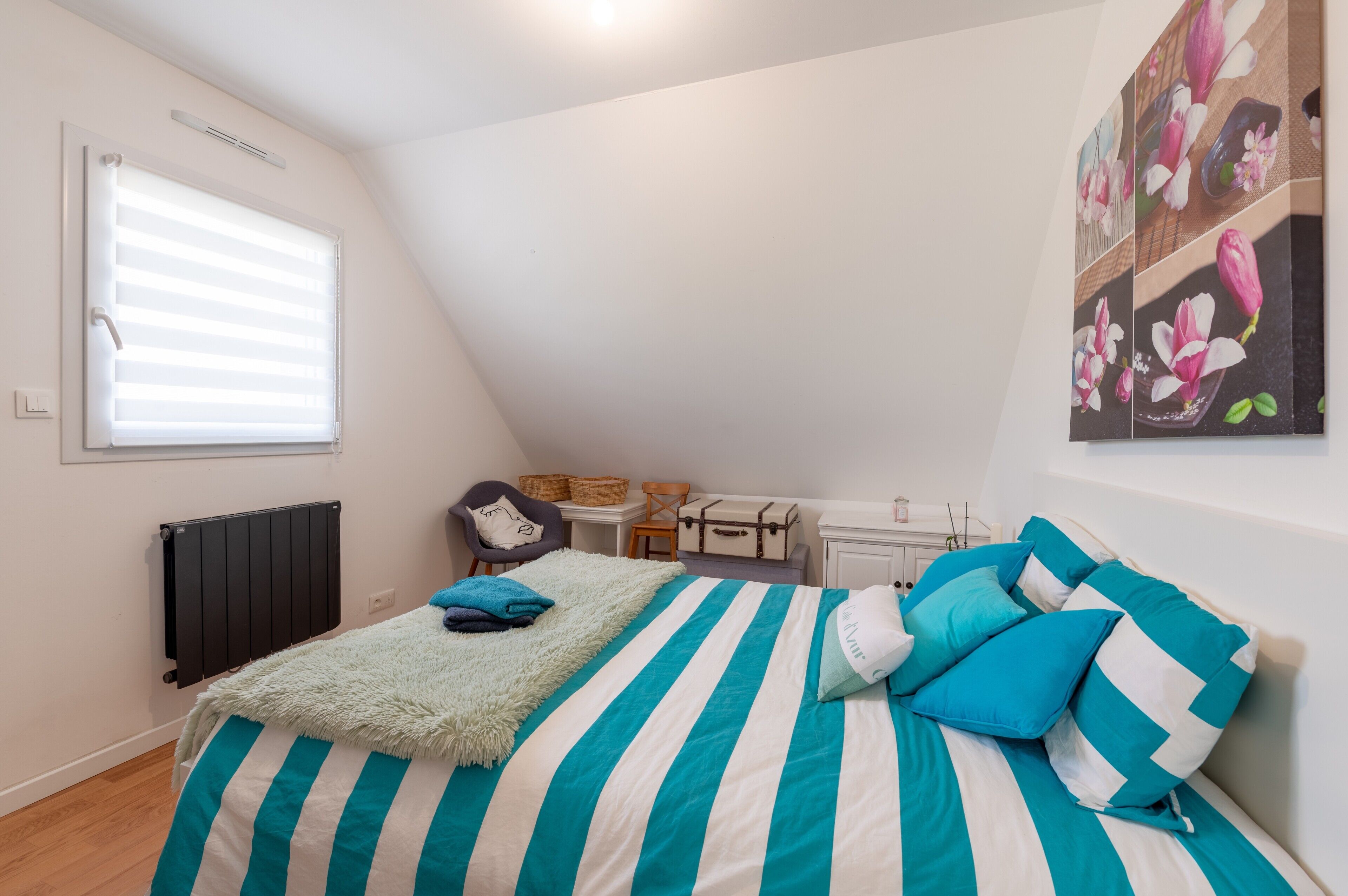 A bedroom with a bed, a radiator, a window with blinds, a chair, a table, and a wall with a floral painting.